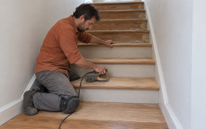 Worker sanding a red oak stair tread by hand in a Baldwin Park townhome after the stair carpet came off, the nosing overhanging each riser.